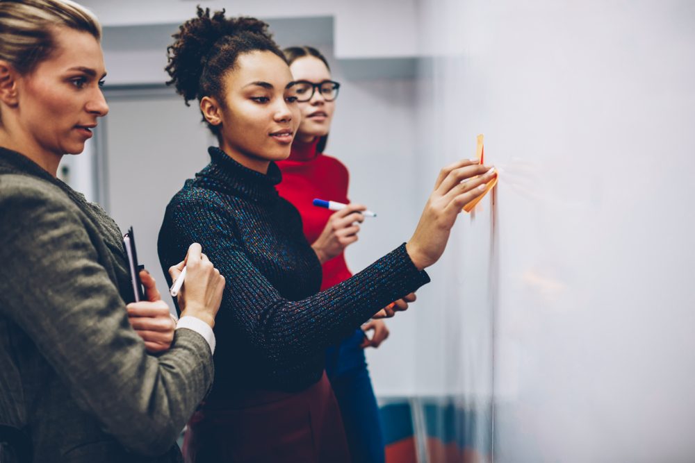Three people write with markers on a whiteboard in an office setting, collaborating and discussing ideas.