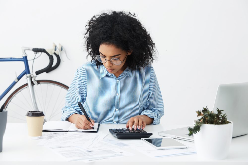 Person writing in a notebook while using a calculator at a desk, surrounded by papers, a laptop, tablet, coffee cup, bicycle, and plant.