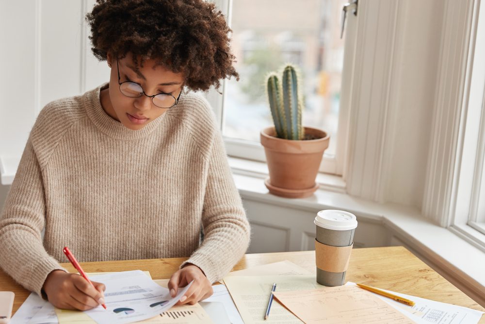 Person writes at a table, surrounded by papers and a coffee cup; a potted cactus sits on the windowsill nearby.