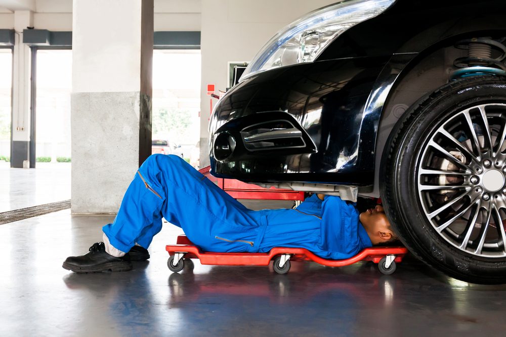 Mechanic in blue uniform lying down and working under car at auto service garage