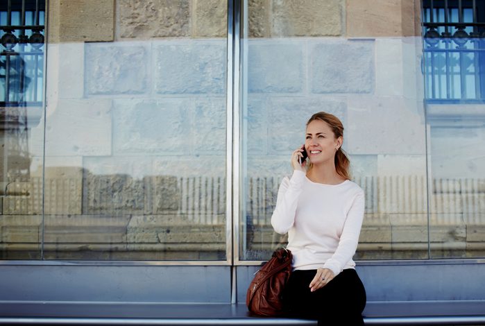 Young beautiful woman talking on mobile phone while waiting public transportation at urban setting station, smiling female having pleasant conversation on cell telephone while sitting on a bus stop