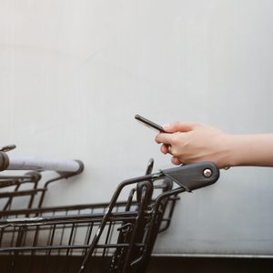 Hand holding a smartphone, typing beside empty shopping carts with a light-colored wall in the background.