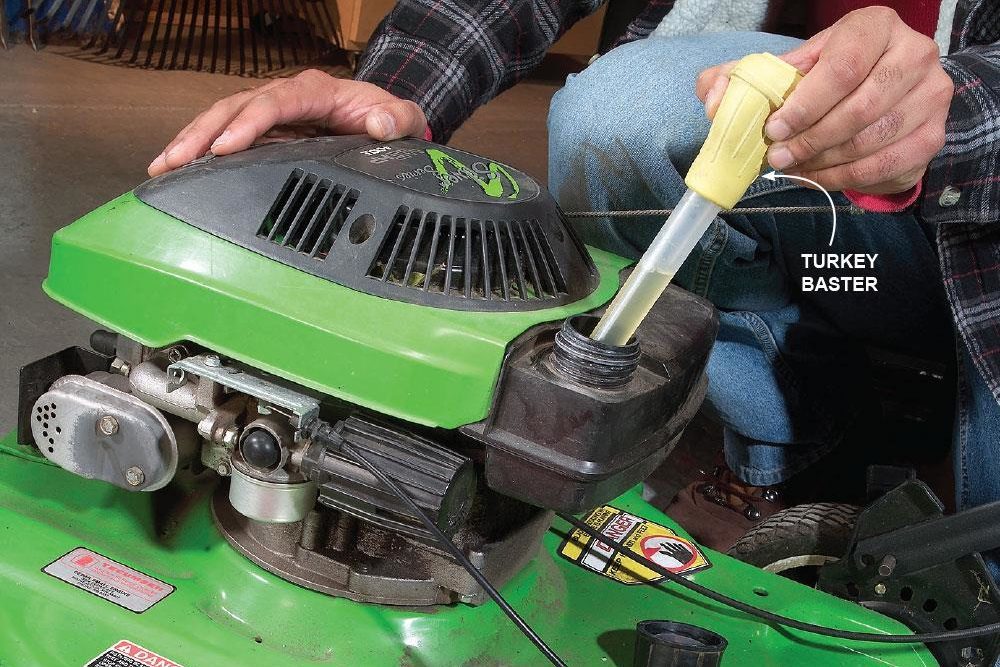 A person uses a turkey baster to extract liquid from a green lawnmower in a garage setting.