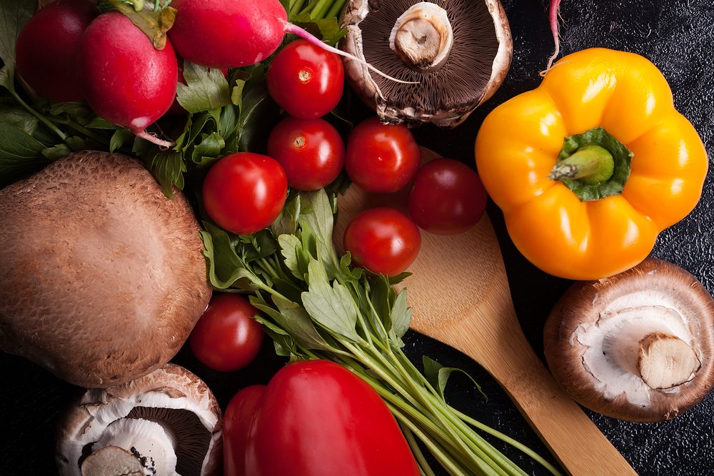 Mushrooms, tomatoes, radishes, and bell peppers rest on a wooden spoon amid leafy greens on a dark textured surface.