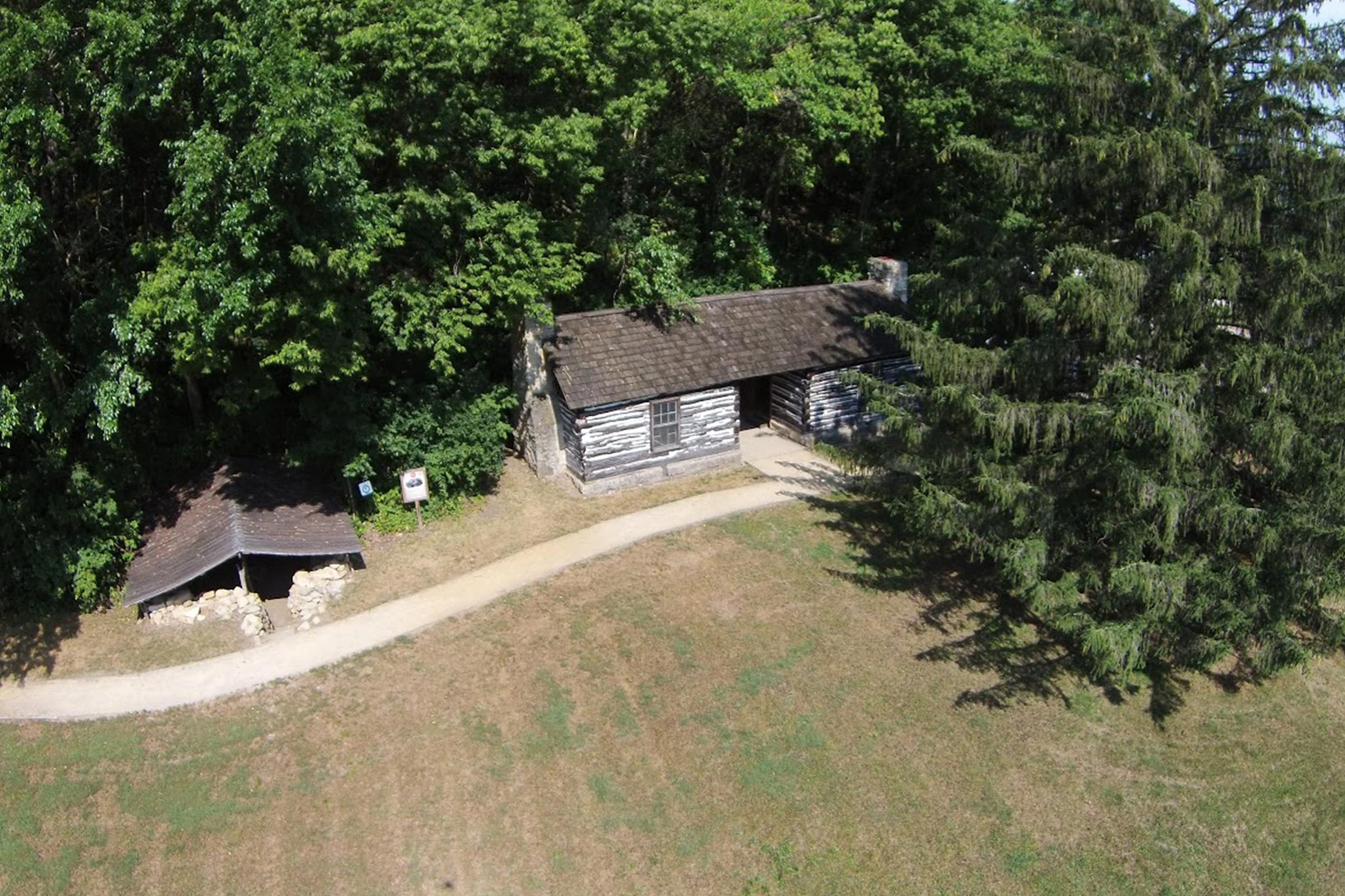 A log cabin sits quietly amidst dense greenery, beside a winding dirt path, with nearby signage and an additional small shelter.