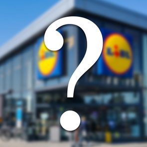 A large question mark obscures the front of a store with logos on a bright, sunny day.