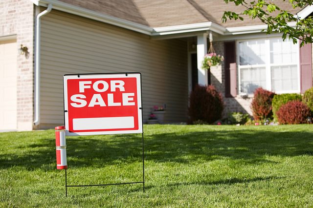 Sign reads "FOR SALE" on a lawn, in front of a house with beige siding, windows, and garden shrubs.