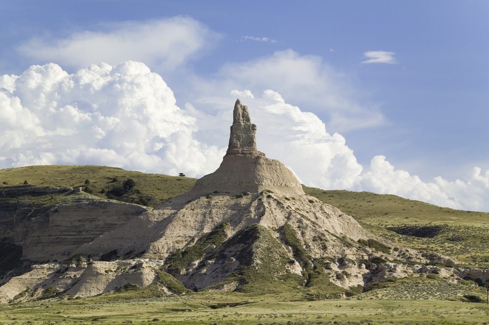A solitary rock formation towers skyward, surrounded by grassy plains and under a sky filled with large, billowy clouds.