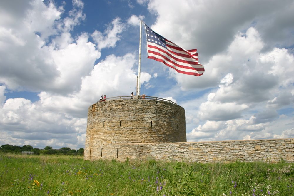 Stone tower displays large American flag, waving under scattered clouds in a grassy field, with people standing on top.