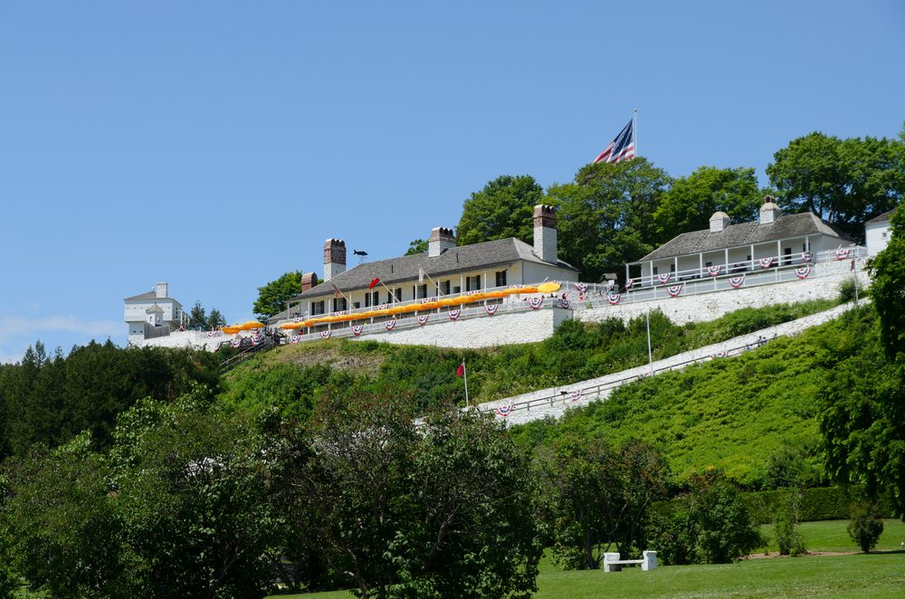 A historic fort, adorned with bunting, sits atop a grassy hill under a clear blue sky with lush trees surrounding it.