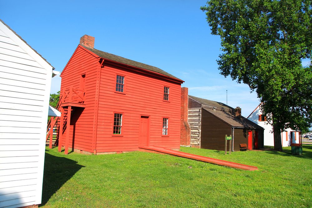 Red wooden house stands on green grass, surrounded by other buildings, under a clear blue sky.