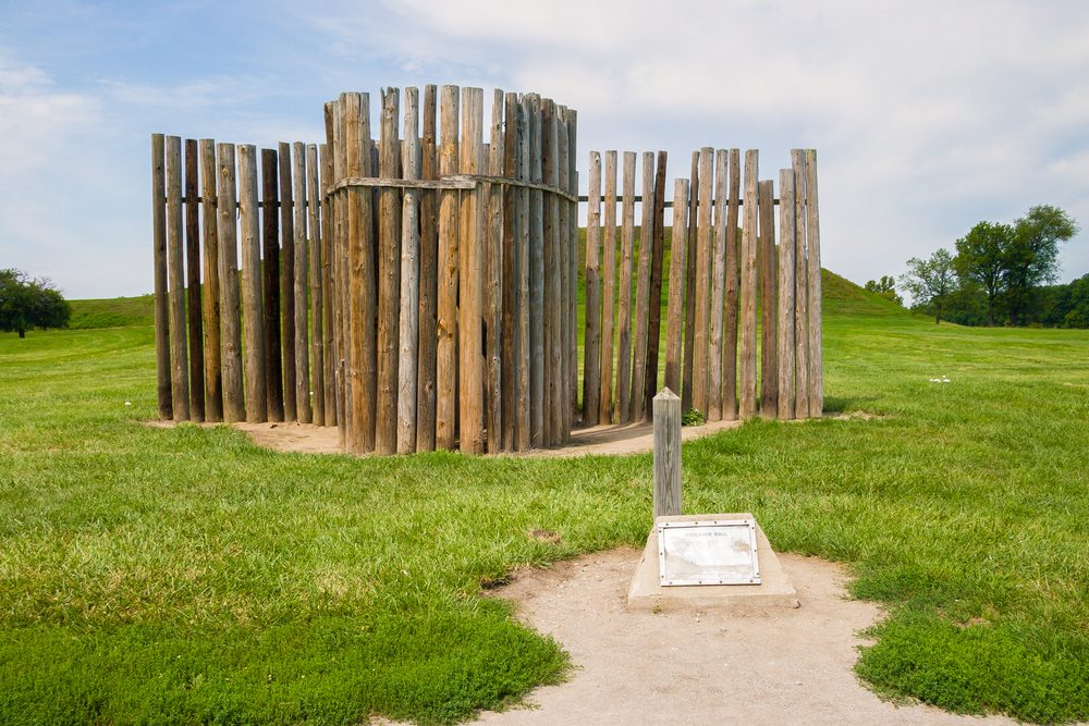 Wooden posts form a circular structure in a grassy field, with a small sign and path in the foreground.