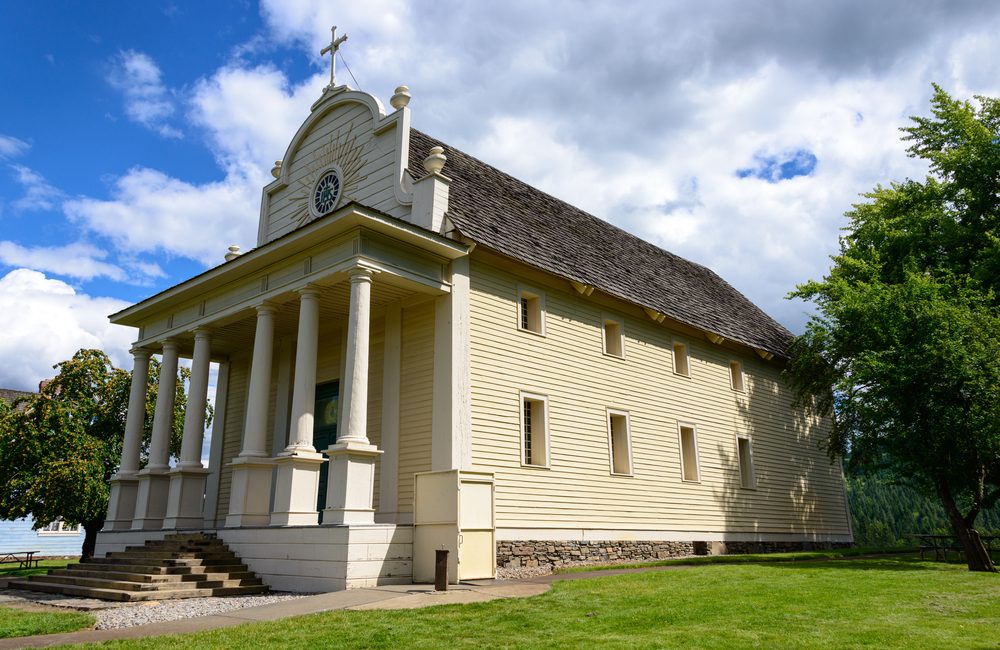 A historic wooden church with columns stands under a cloudy sky, surrounded by green grass and trees.