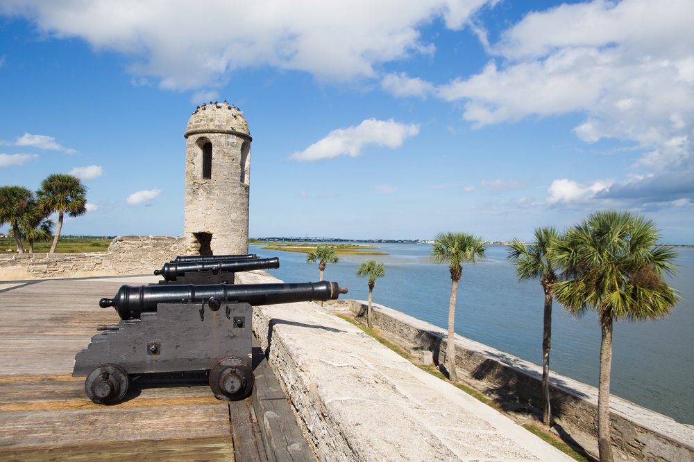 Cannons point toward water, positioned on a historical stone fort wall. Palmetto trees and a watchtower accentuate the coastal landscape under scattered clouds.