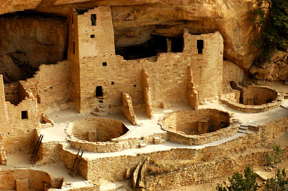 Ancient stone structure stands nestled in a cave, surrounded by circular pits and ladders, with rugged rock formations overhead.