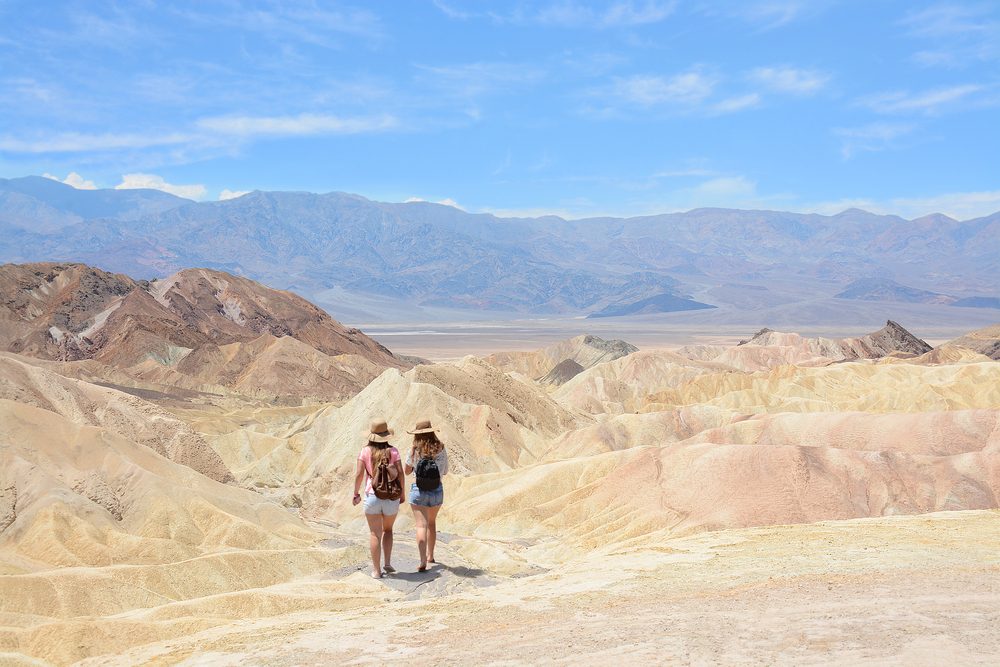 Two individuals walk on rugged, colorful desert hills under a clear blue sky, surrounded by distant mountains.