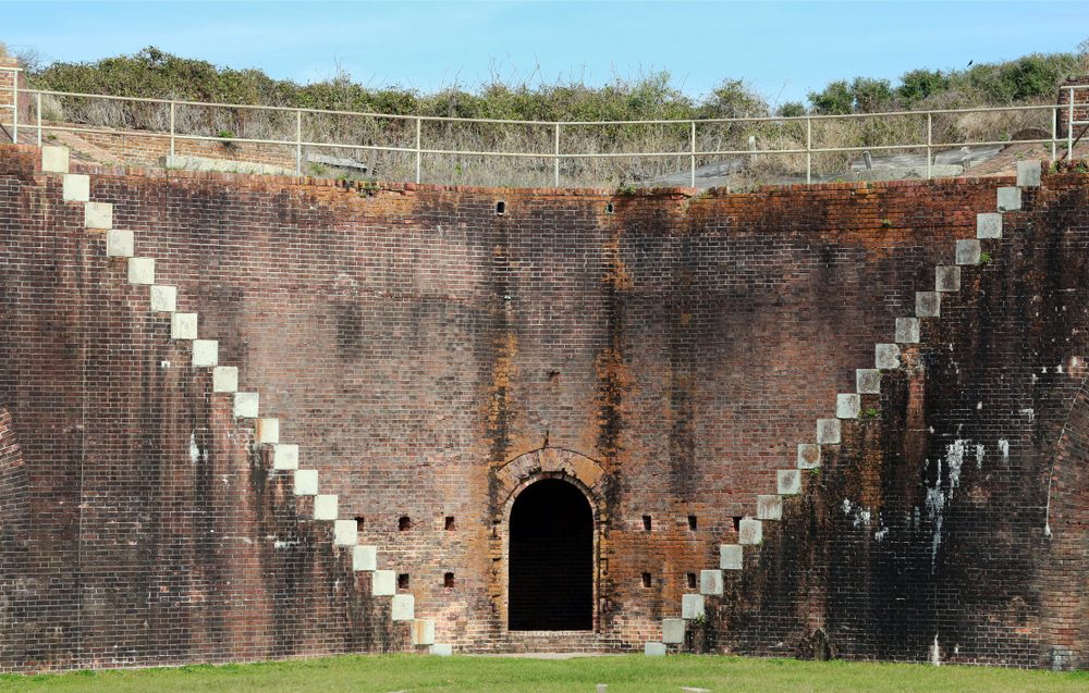 Brick fort wall features steps leading to a central arched door; surrounded by green grass and topped with a railing, under clear sky.