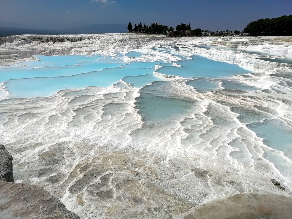 Terraces of bright blue water filled limestone pools glimmer under sunlight, bordered by trees in the background.