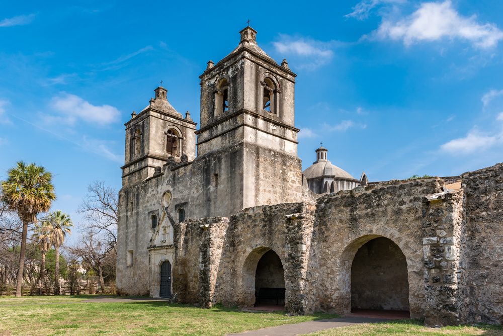 Stone church stands, weathered facade, under a bright blue sky; palm trees and grass fill the surrounding area.