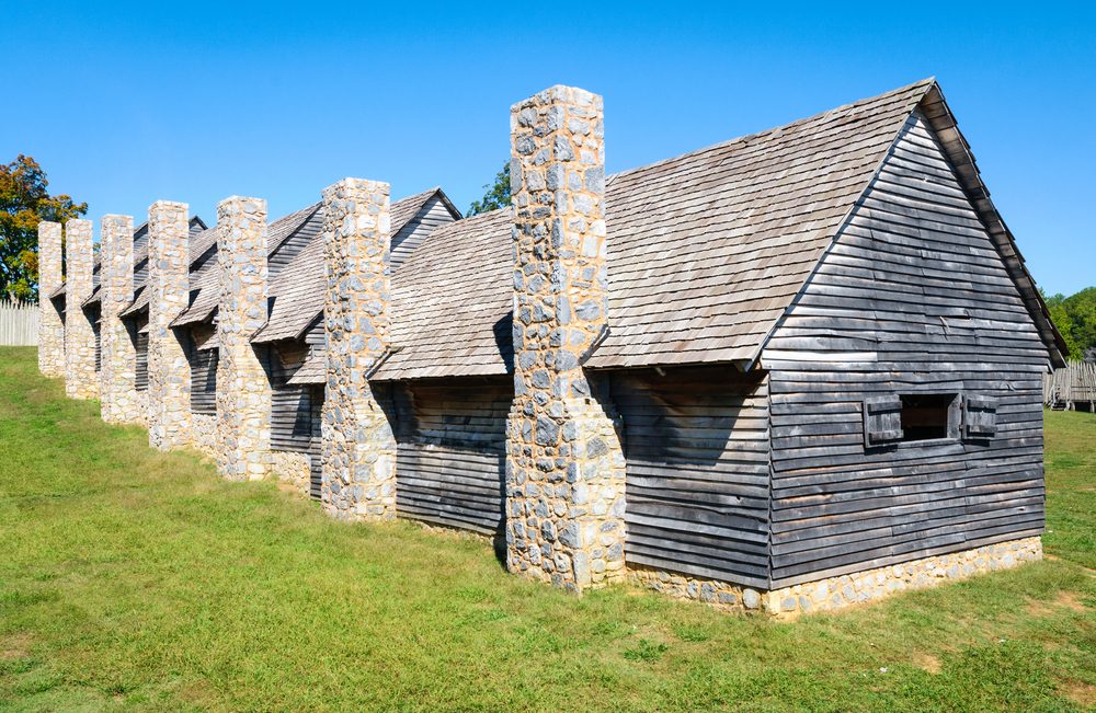 Wooden cabins feature stone chimneys in a grassy historical settlement under a clear blue sky.