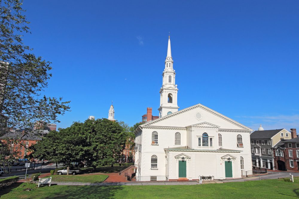 White church stands prominently, surrounded by lush trees and historic buildings under a clear blue sky.