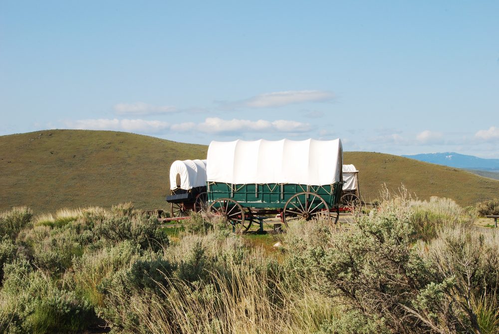 Covered wagons stand still on grassy plains, surrounded by rolling hills under a clear blue sky.