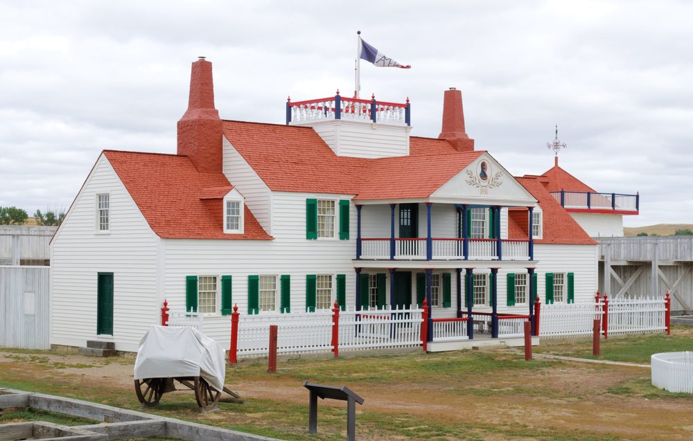 Building stands with red roof, four chimneys, and a second-floor balcony; surrounded by a white fence on a cloudy day. A cart is nearby.