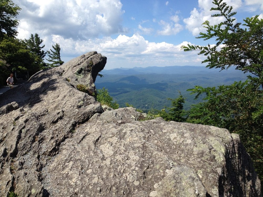 Rocky outcrop juts over a forested valley, surrounded by trees under a bright, partly cloudy sky.