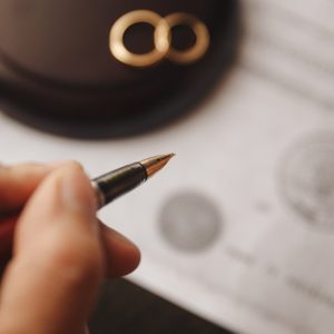 Hand holds pen, signing document on a table, with two wedding rings and a gavel nearby.