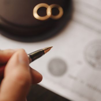 Hand holds pen, signing document on a table, with two wedding rings and a gavel nearby.