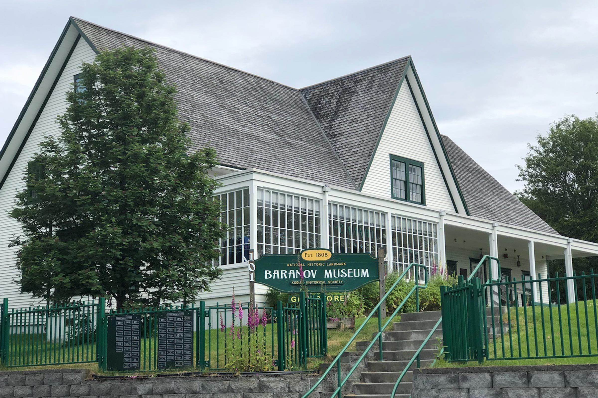 Building labeled "Baranov Museum" stands. Stone steps lead up. Green fence and trees surround the wooden, historic structure. Overcast sky overhead.