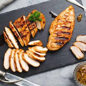 Grilled chicken slices arranged on a slate board with mustard sauce, antique cutlery, and a napkin nearby.