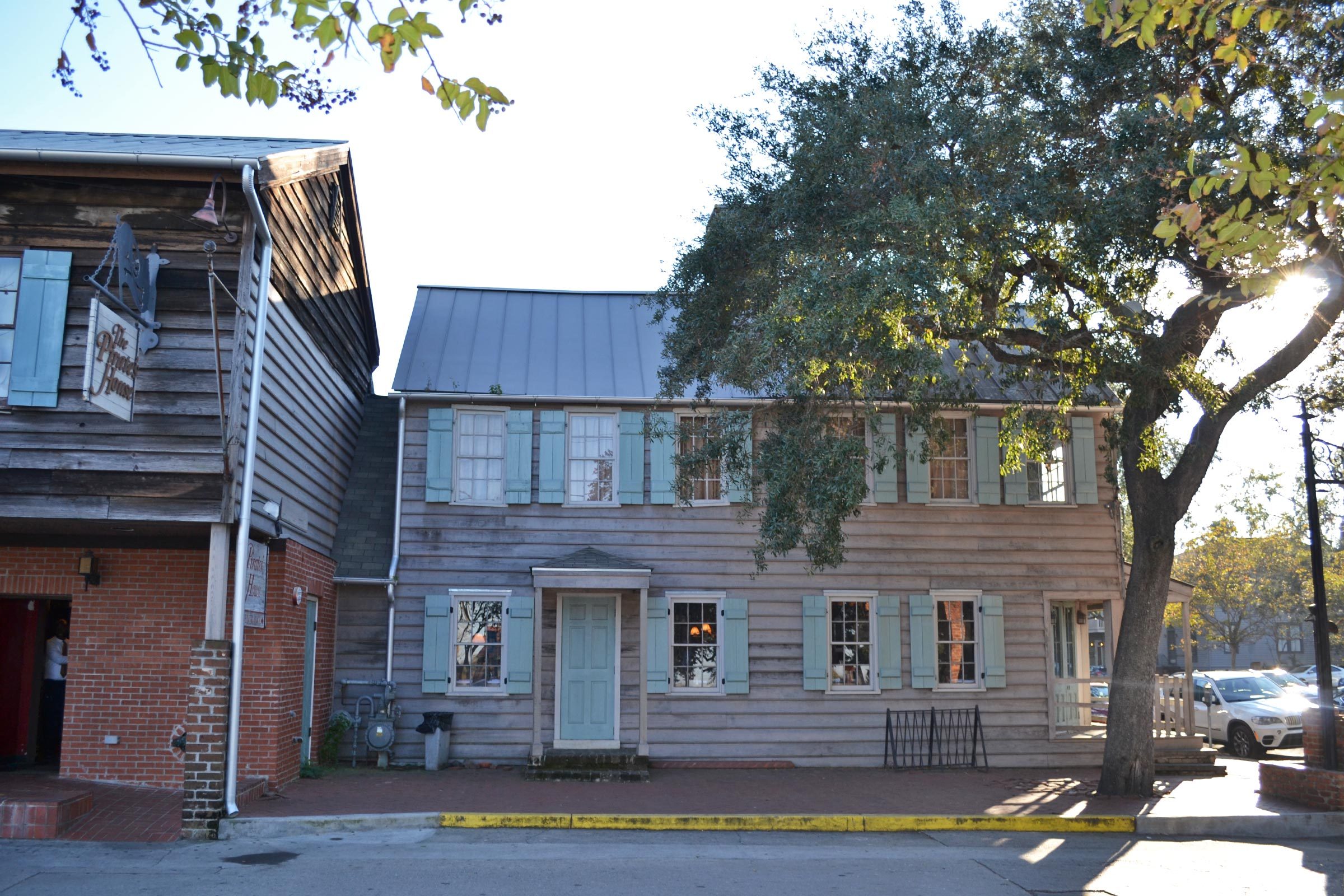 A wooden building with blue shutters stands in sunlight beside a tree and adjacent brick building under a clear sky. Sign reads â€œThe Printer.â€