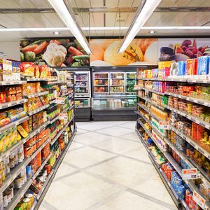 Grocery shelves display various food items in a brightly lit supermarket aisle with a colorful produce mural overhead and refrigerators at the back.