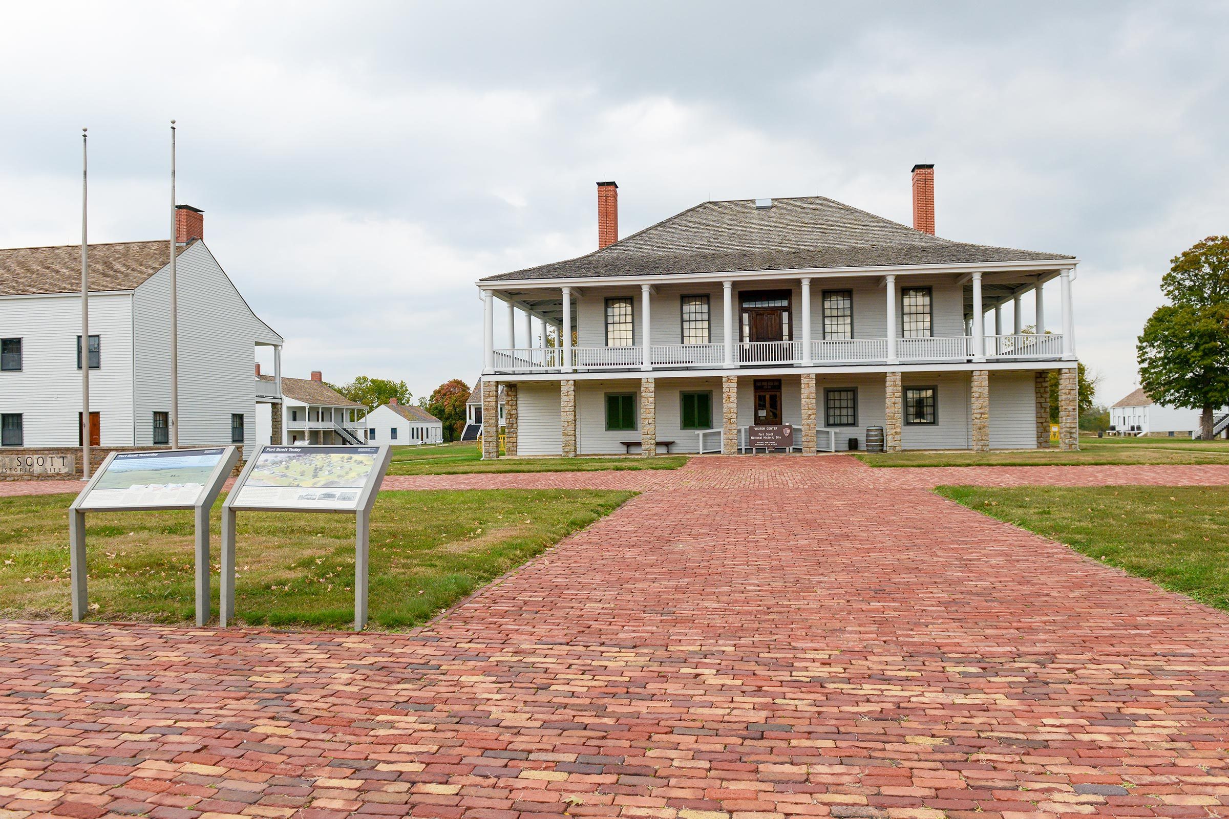 A two-story, historical building stands with a wraparound porch, surrounded by grassy lawns and brick pathways under a cloudy sky. Nearby are informational plaques.