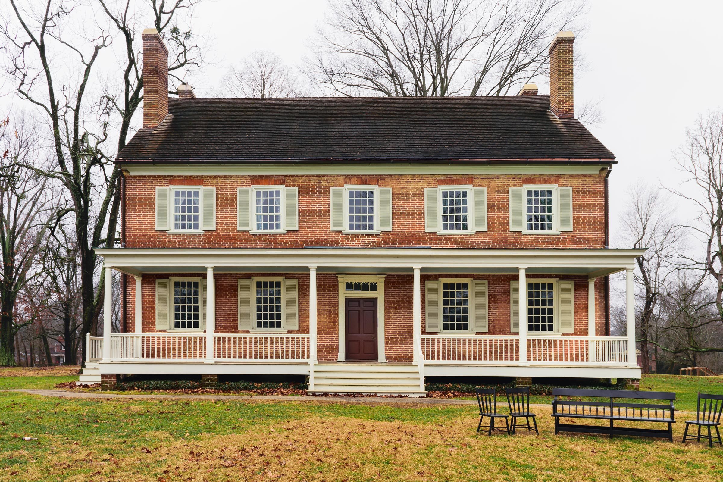 Brick house with shutters stands still, surrounded by leafless trees and lawn furniture, set in a grassy landscape with a cloudy sky.