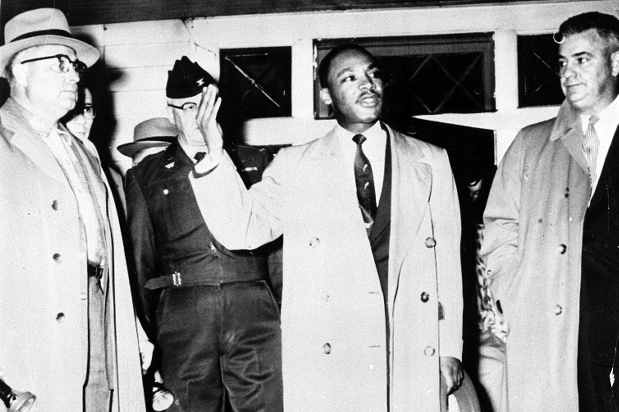 A man gestures while speaking; surrounded by men in coats and a uniformed officer, indoors at night.