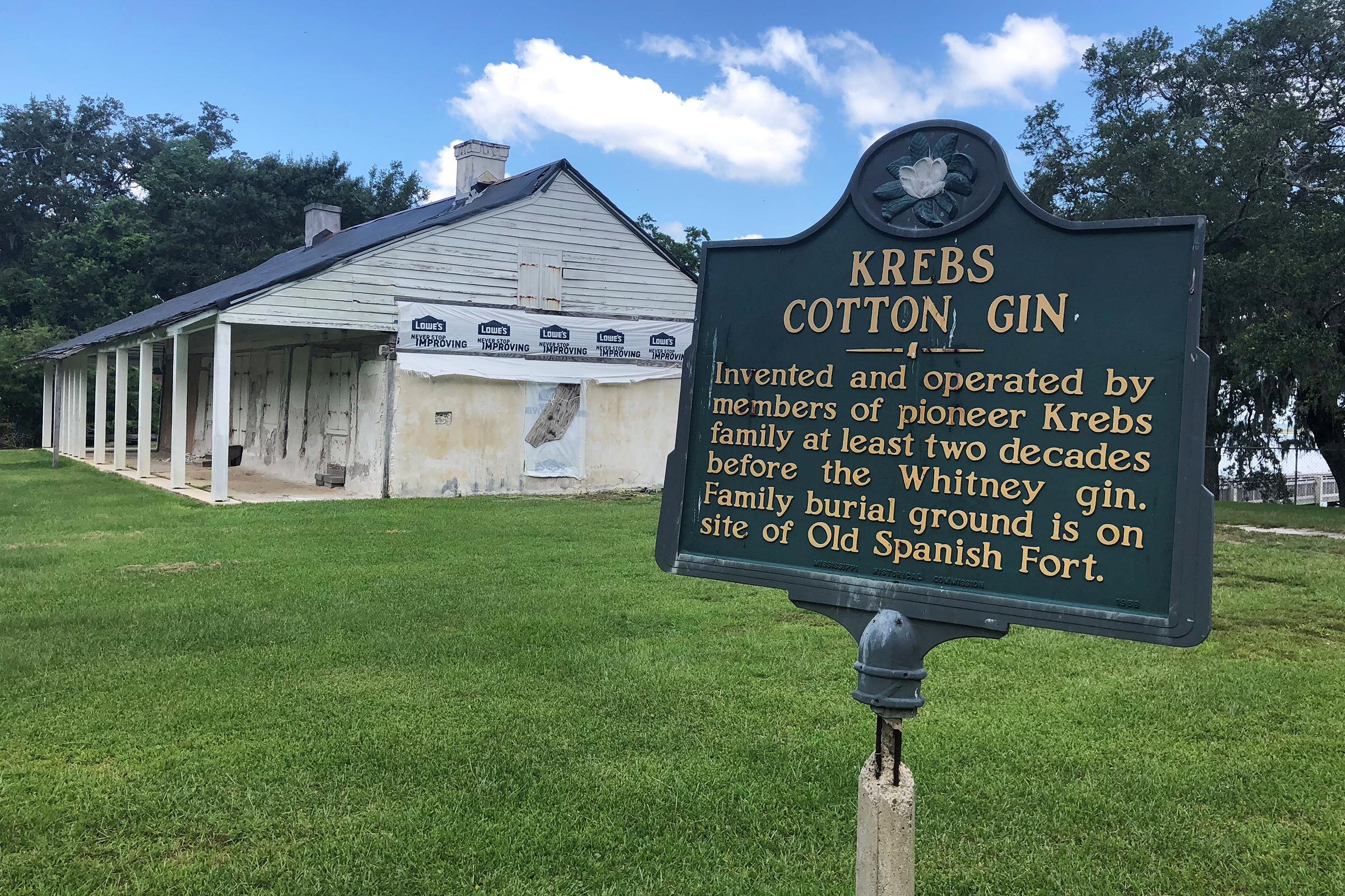 Sign describes Krebs Cotton Gin history; stands before an old white wooden structure under renovation, surrounded by grass and trees under a blue sky.