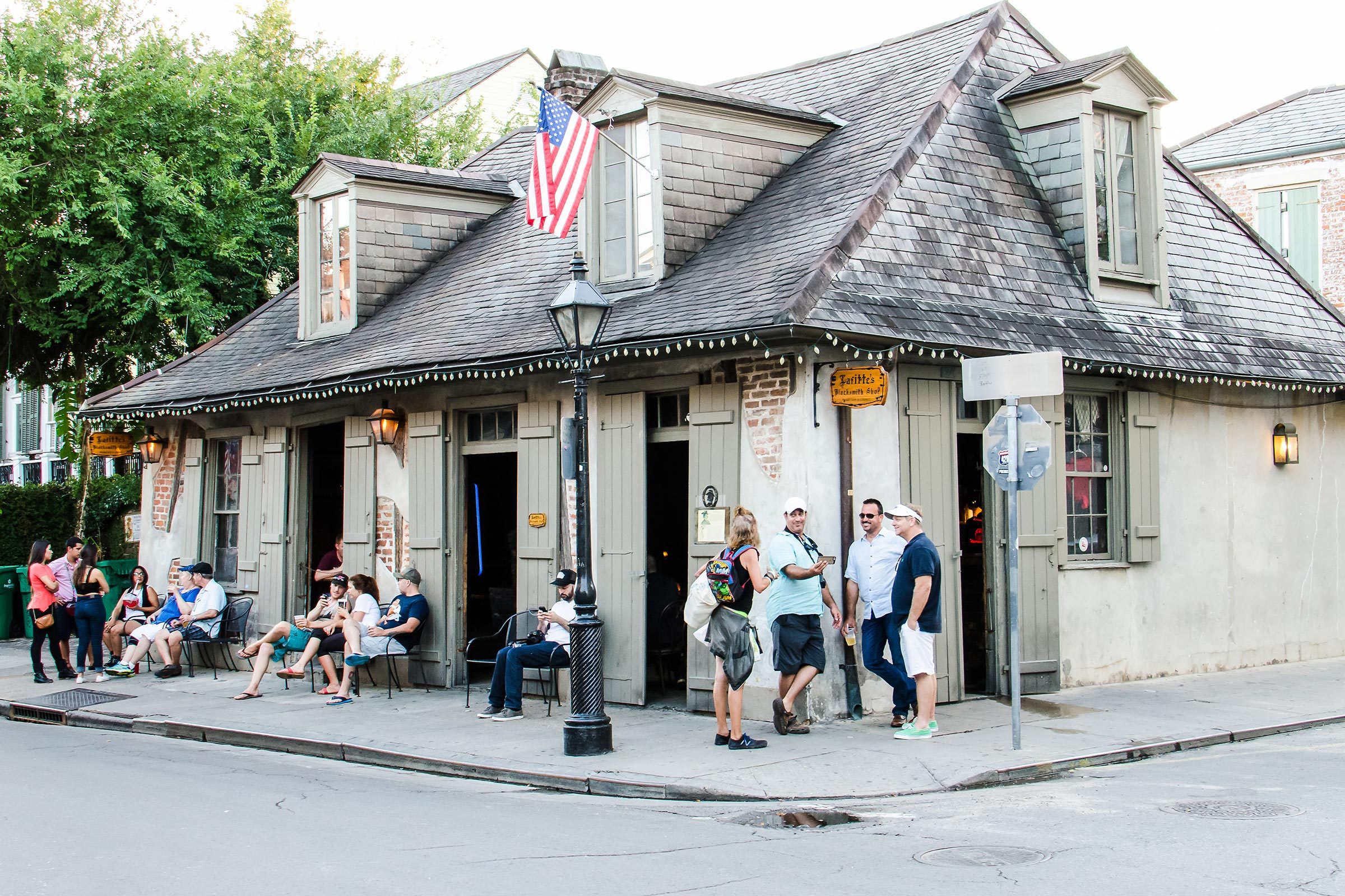 Tavern with American flag, people sitting and standing outside; historical architecture on a street corner under trees.