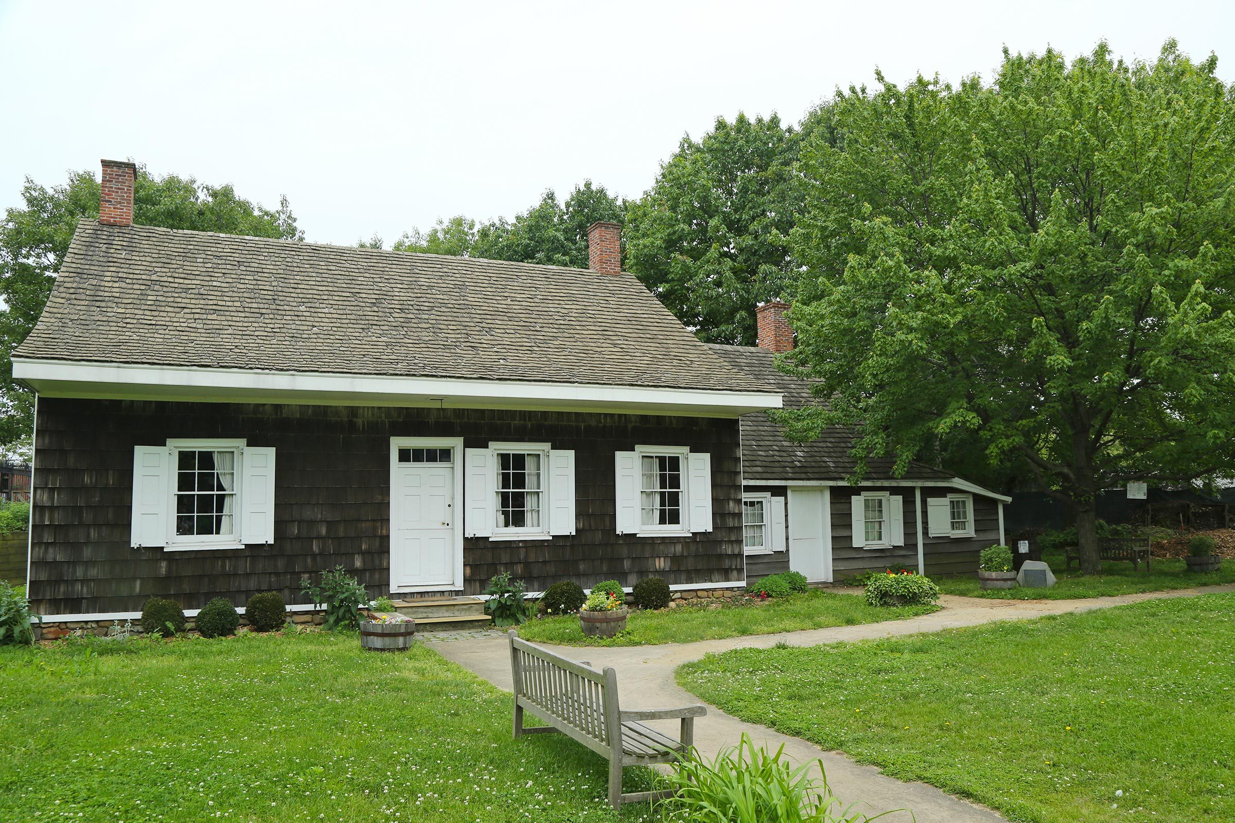 A wooden house sits quietly amidst lush greenery, featuring white shutters and a bench on the lawn.