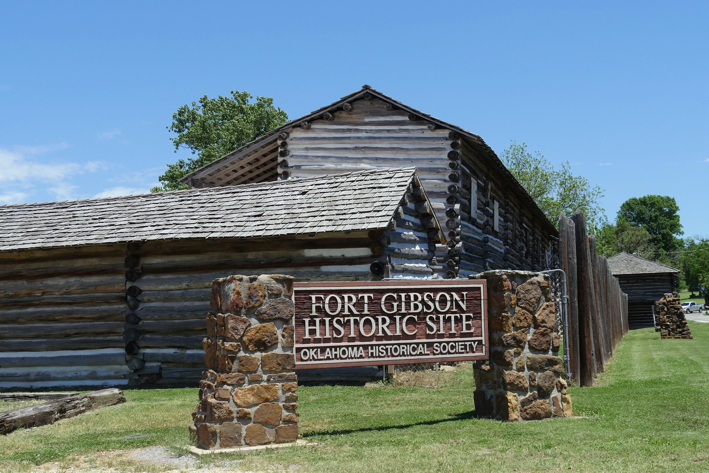 Sign displays "Fort Gibson Historic Site, Oklahoma Historical Society" beside a log building, under a clear blue sky with surrounding greenery.