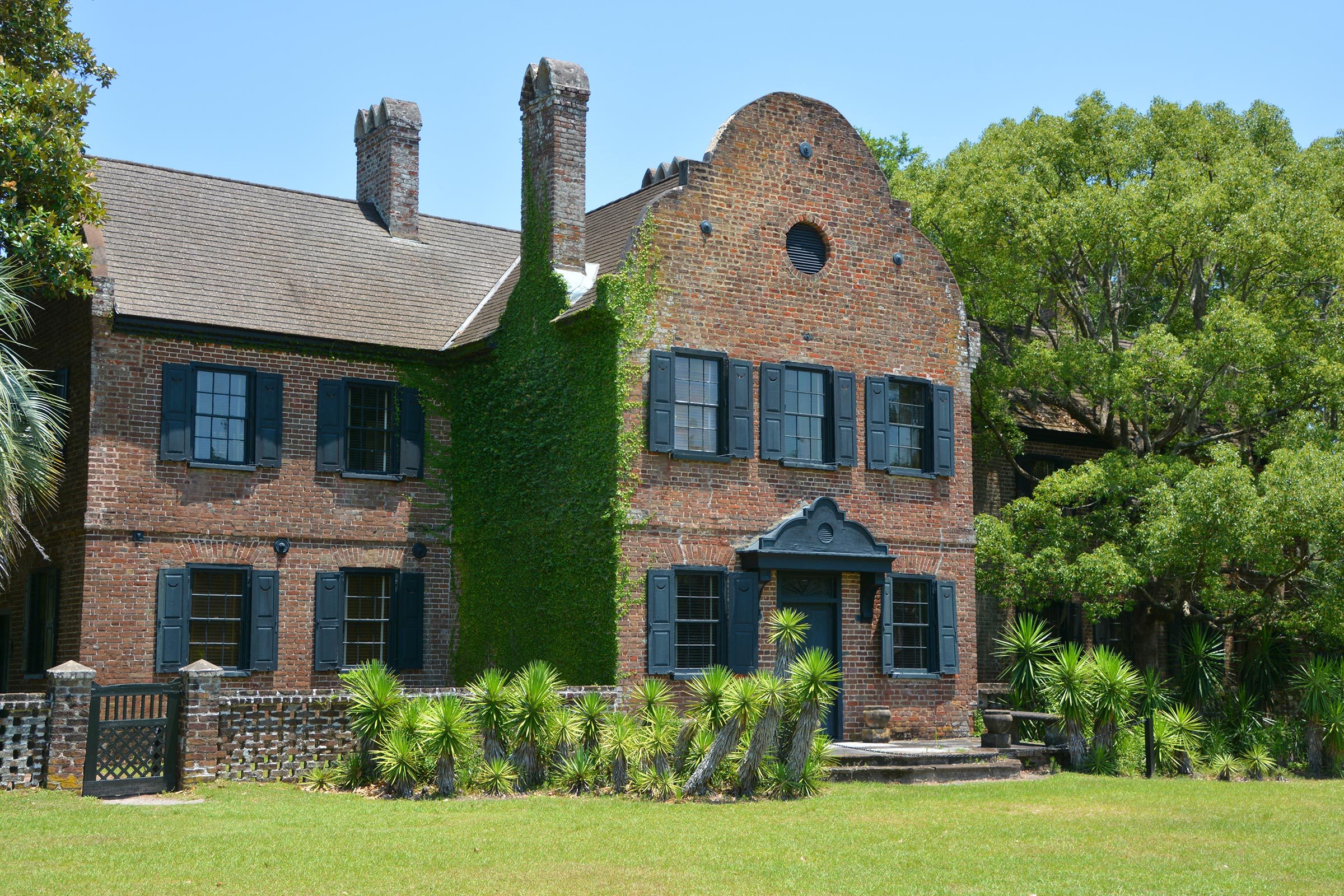 Brick house stands covered in ivy, surrounded by trees and grass, under a clear blue sky.