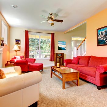 Bright living room features red furniture, ceiling fan, and paintings, with a large window and fireplace, creating a cozy, inviting atmosphere.