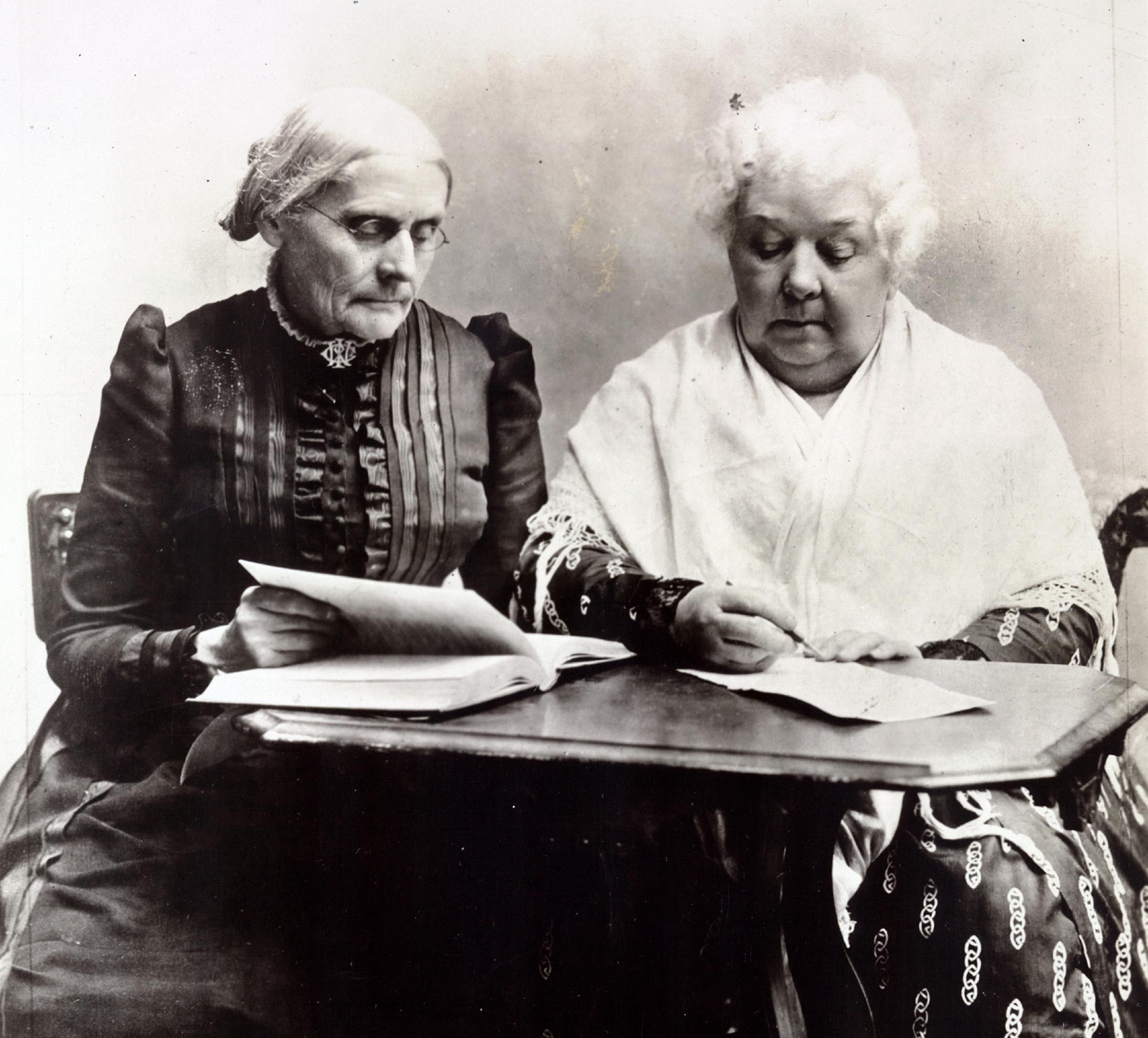 Two elderly women sit at a table; one reads a book, the other writes on paper, in a formal setting.