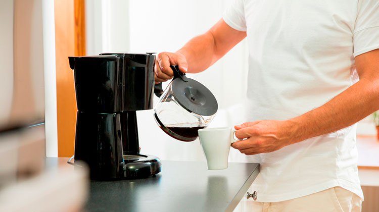 Person pours coffee from a pot into a white mug, standing next to a black coffee maker in a kitchen setting.