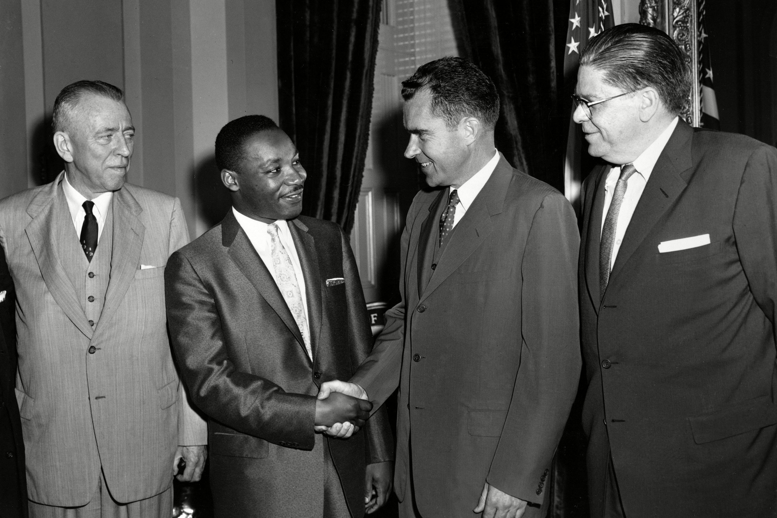 Four men stand in suits; two are shaking hands. They are indoors with curtains and an American flag in the background.