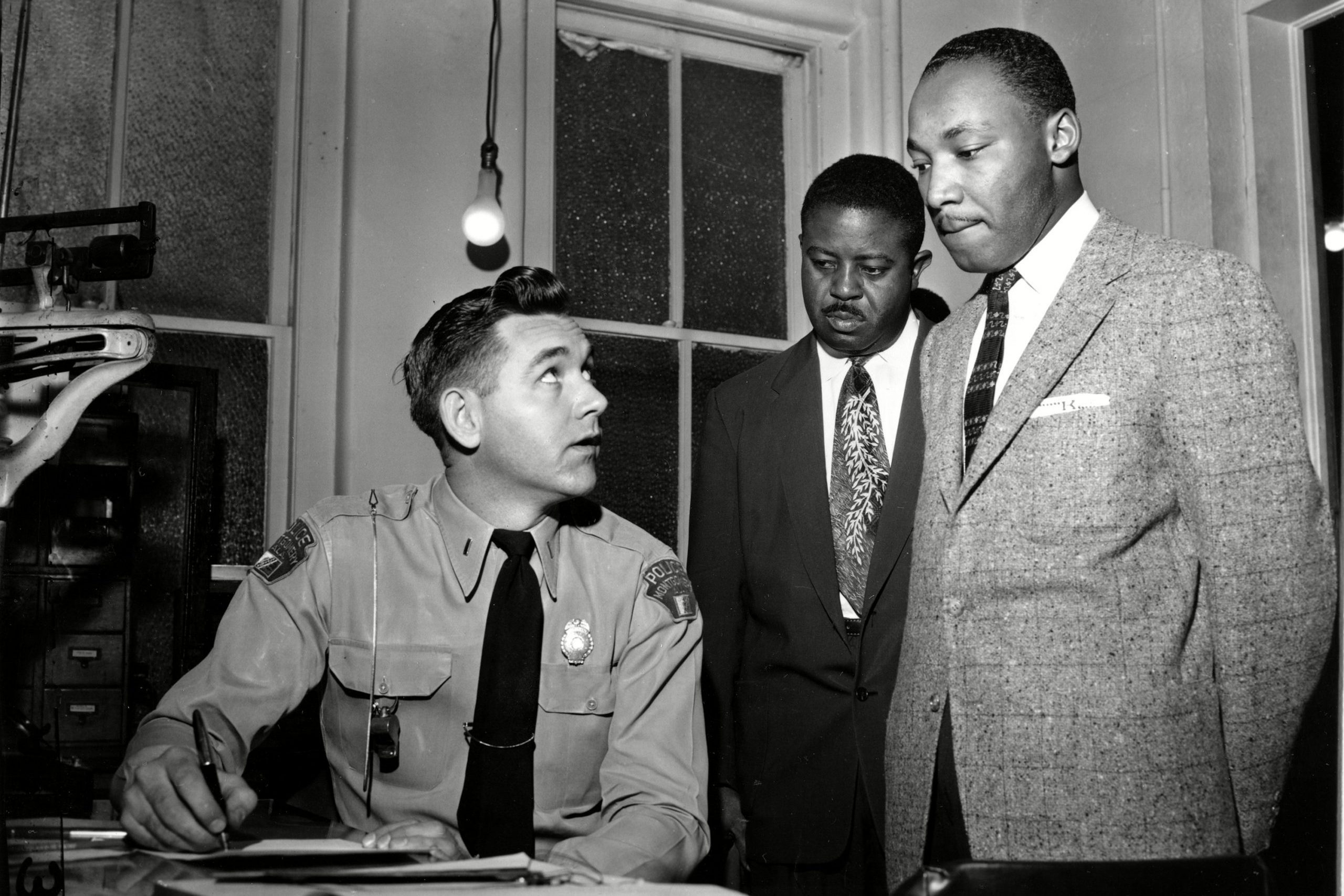 A police officer writes at a desk while two men in suits look on in an office with large windows and a hanging light bulb.