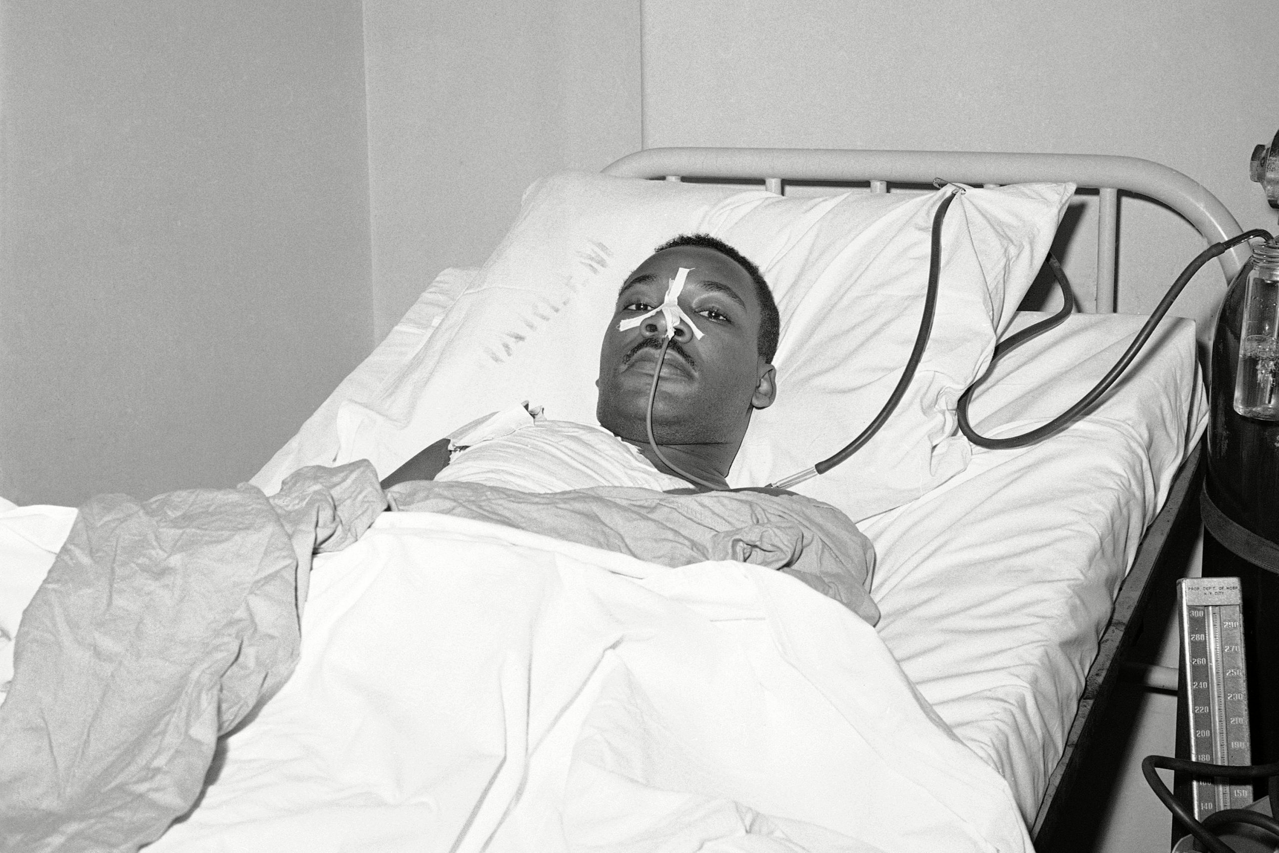 A man lies in a hospital bed with bandages and tubes, in a clinical setting.