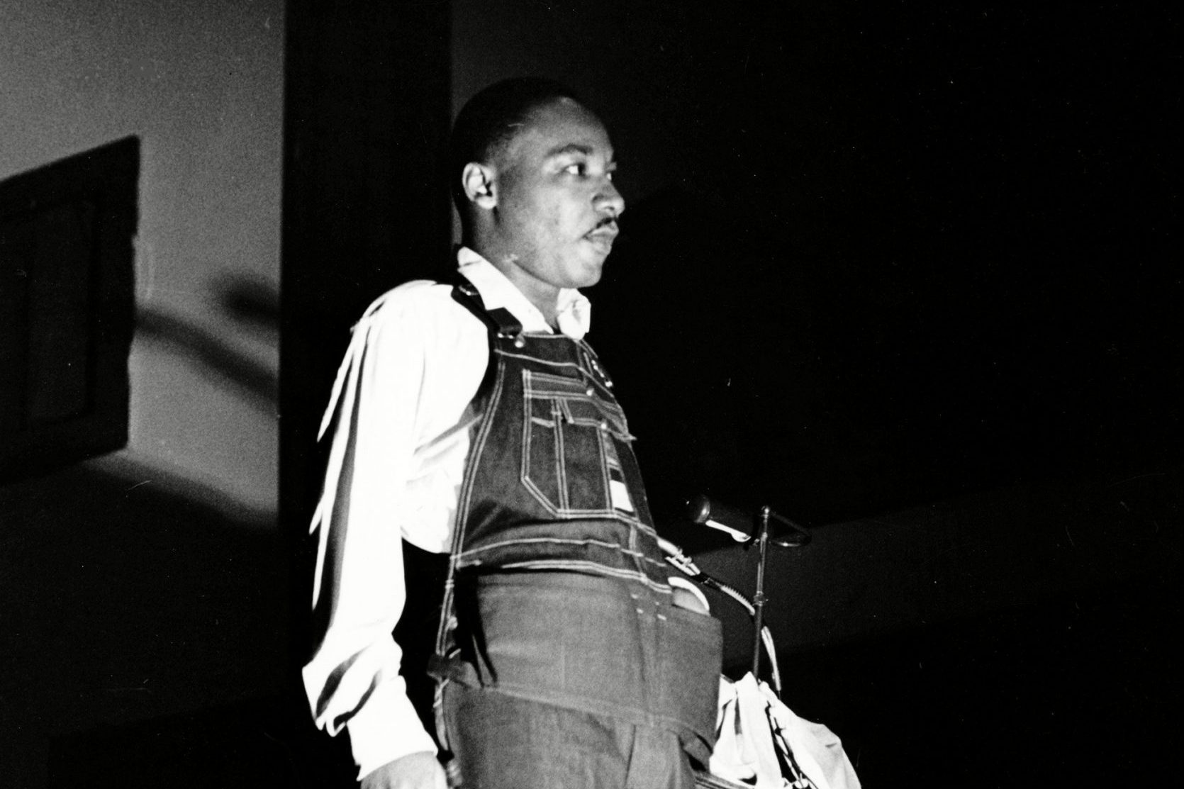 Man standing, wearing denim overalls and a white shirt, looking forward in a dimly lit room with a microphone stand nearby.