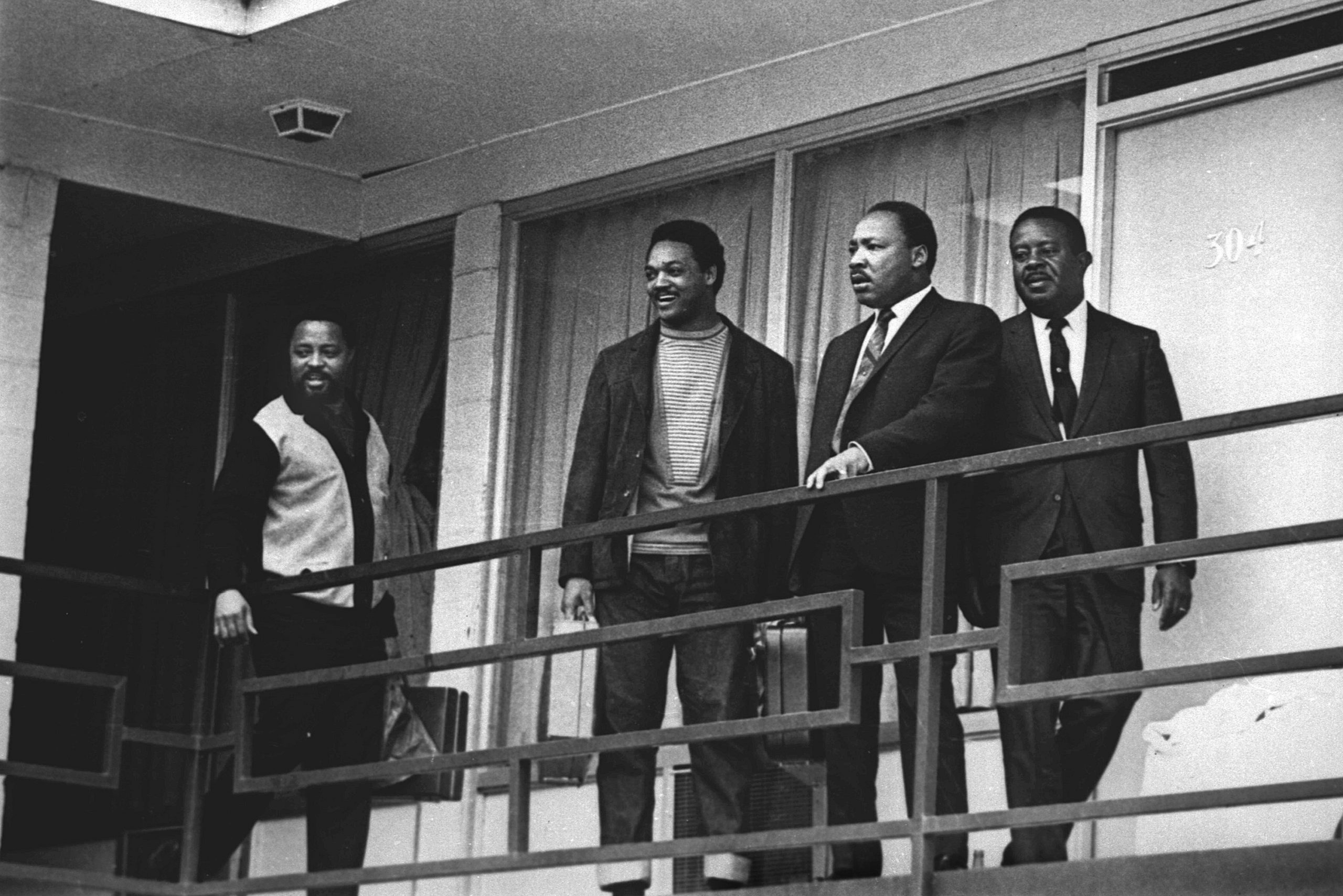 Four men stand on a balcony, engaged in conversation, outside a building with a door labeled "304."